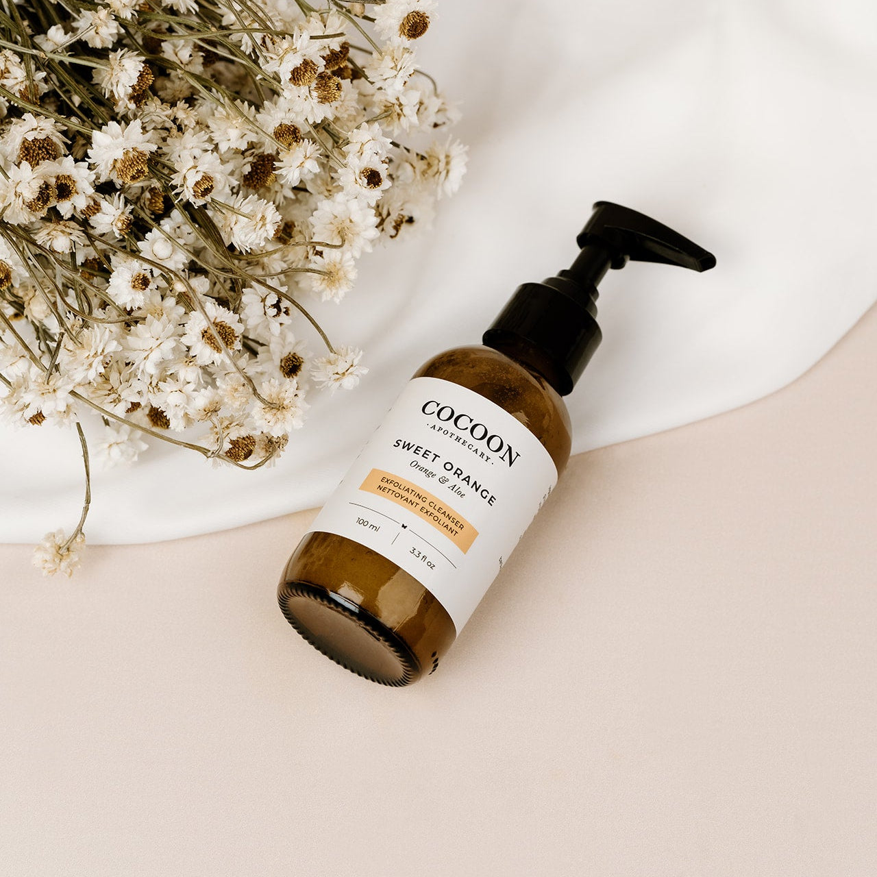 Cocoon Sweet Orange bottle of facial cleanser on a beige surface with white flowers in the background