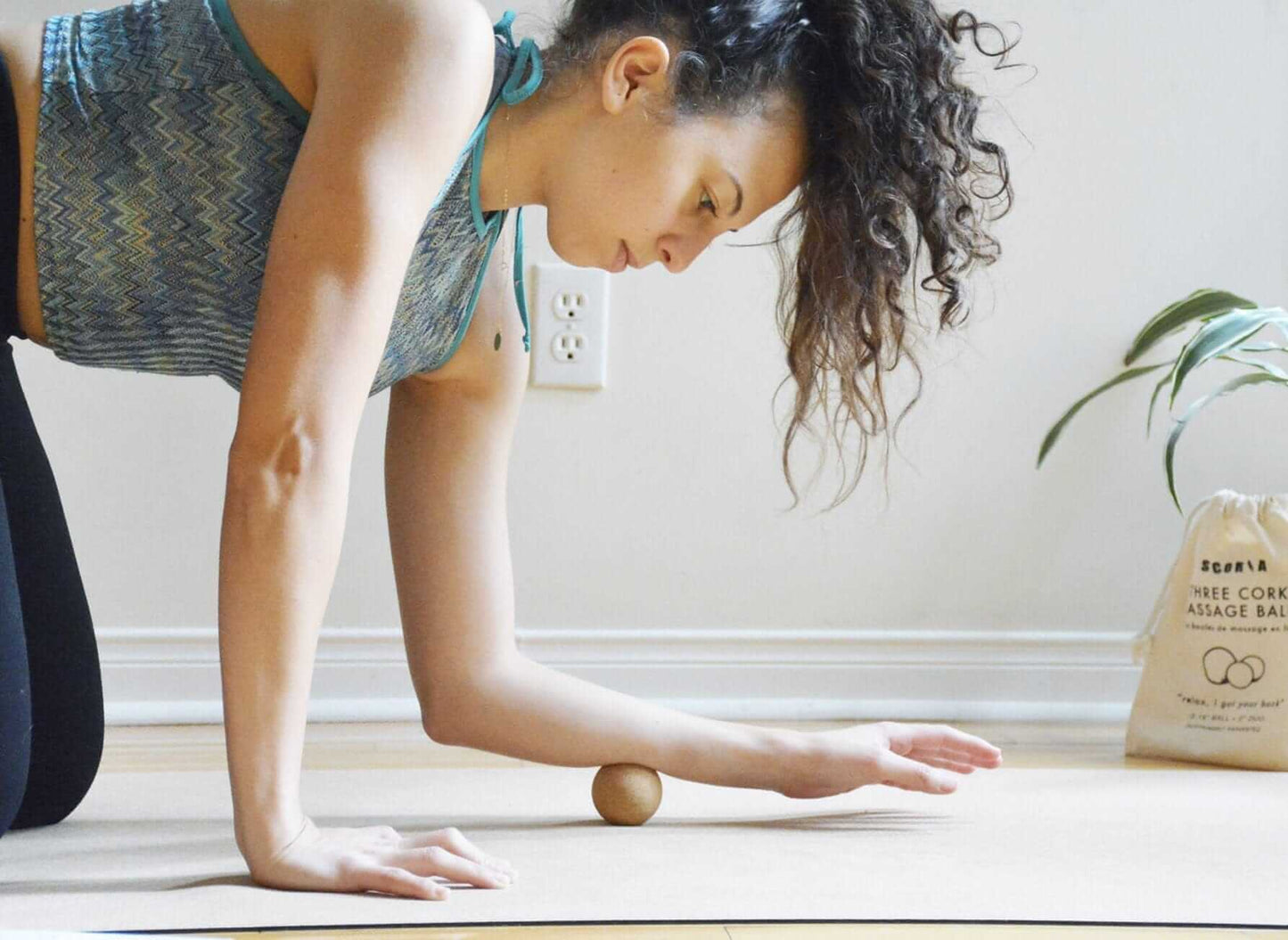 Woman in a plank position with a massage ball on a wooden floor, next to a bag of tea.