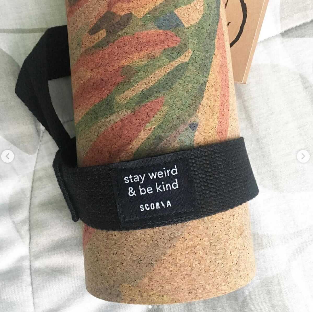 Cork yoga mat with a black strap and 'stay weird & be kind' message on a neutral background