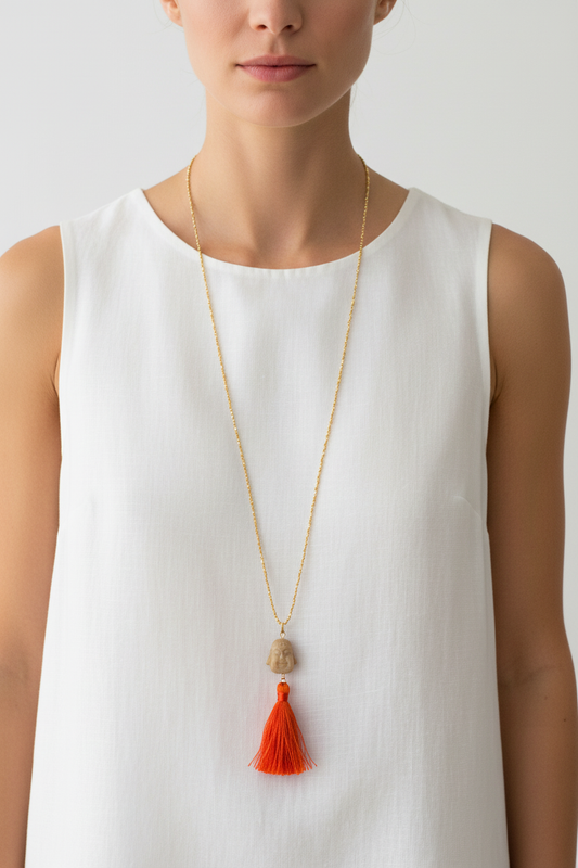 Long gold seed bead necklace with natural stone carved Buddha head pendant and vibrant orange and red silk tassel worn by a woman — handmade in India for boho, yoga, and spiritual style