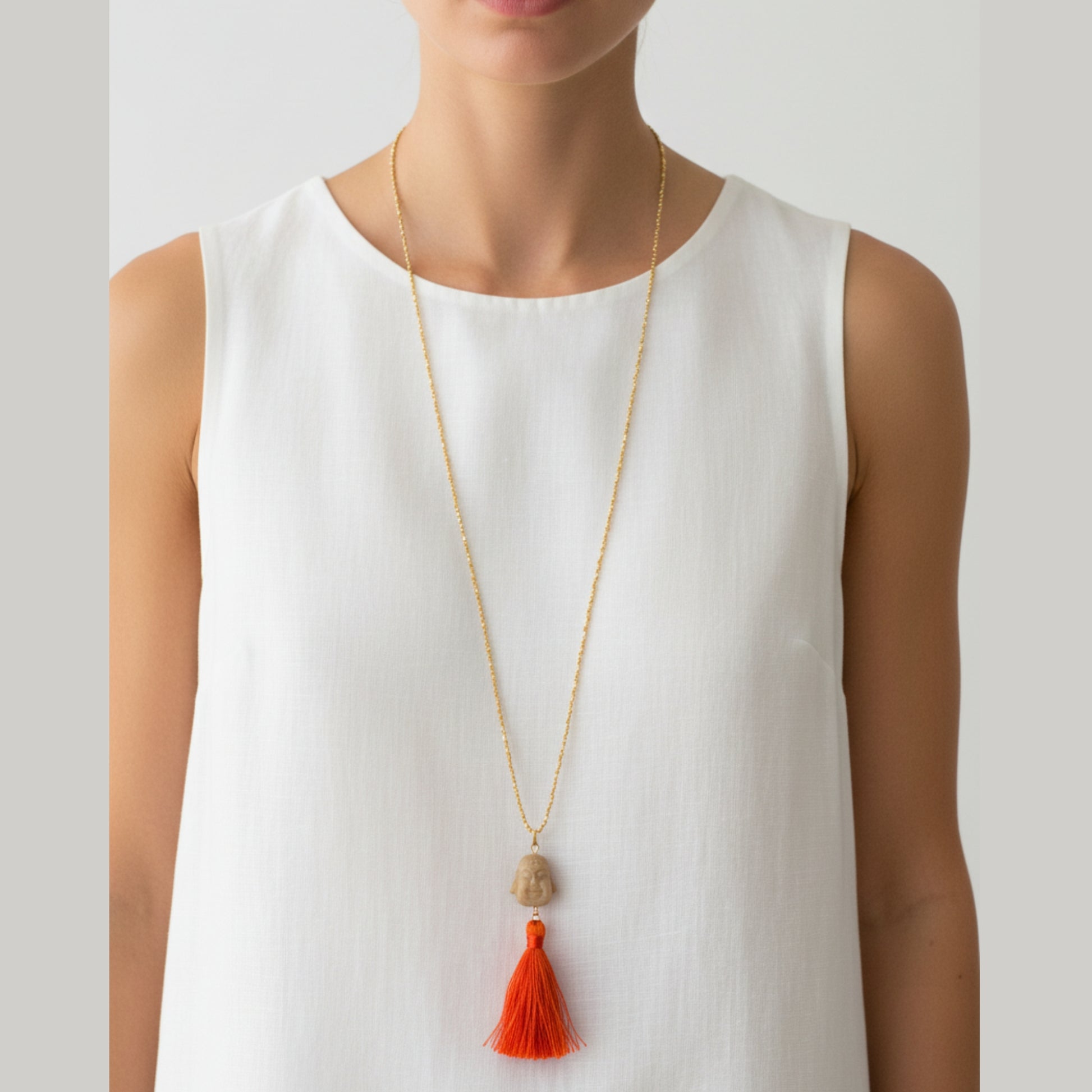 Long gold seed bead necklace with natural stone carved Buddha head pendant and vibrant orange and red silk tassel worn by a woman — handmade in India for boho, yoga, and spiritual style