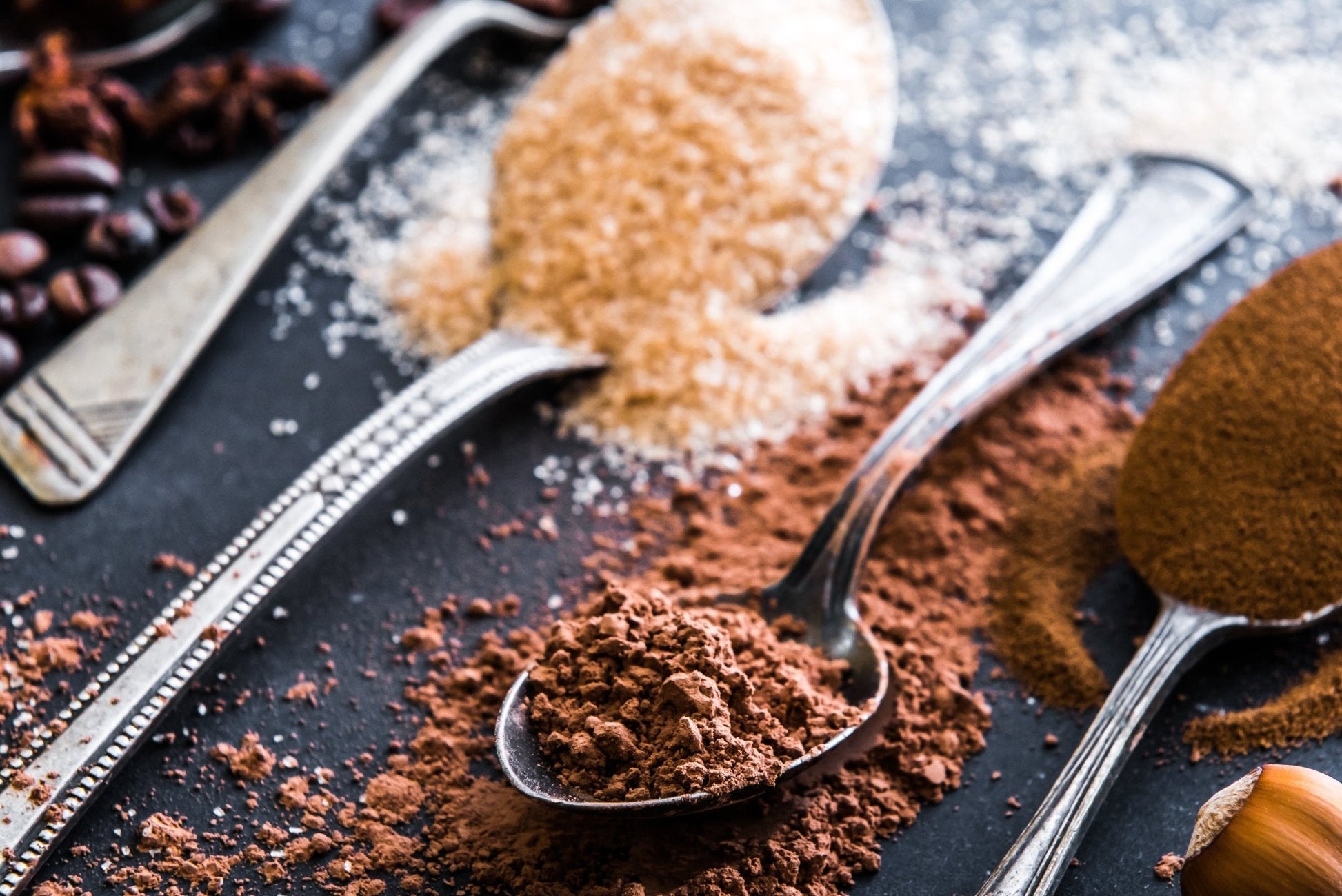 Spoons filled with ground coffee, cocoa powder, and brown sugar on a dark surface.