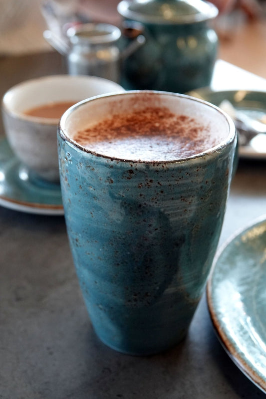 Blue clay pottery cup filled with hot chocolate on a table with other coffee cups and a spoon.