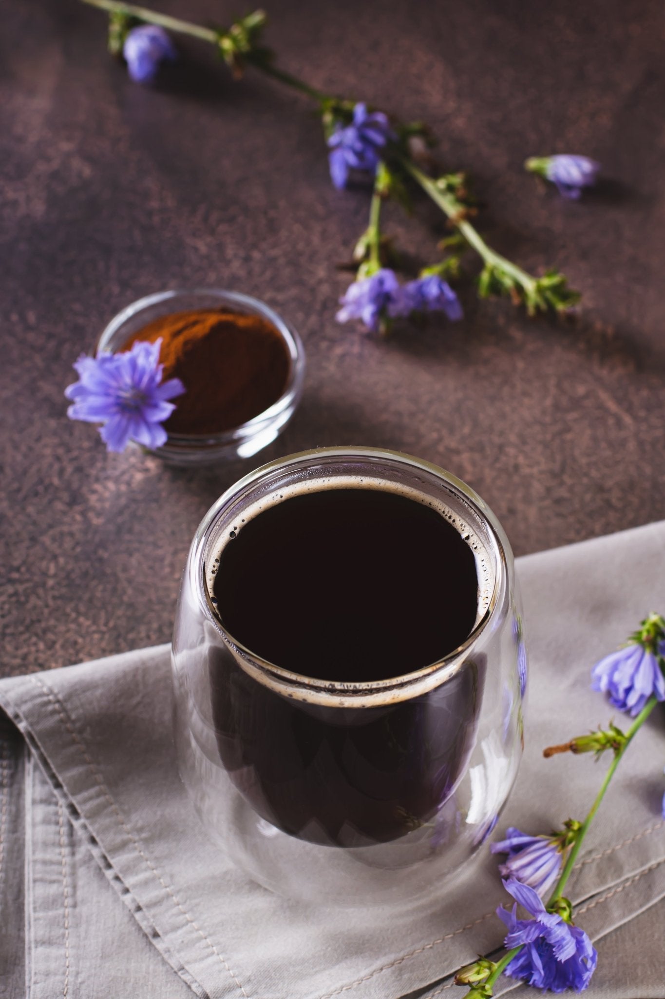 Glass jar of dark liquid with a small container of liquid and purple flowers on a brown surface.
