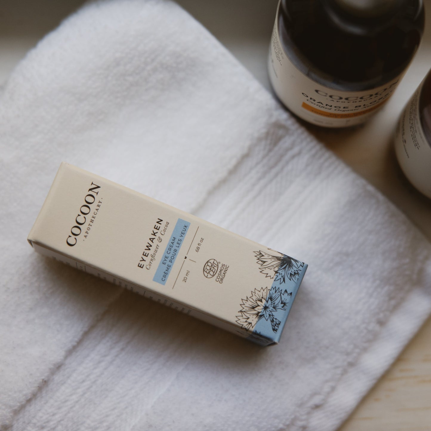Cocoon skincare product 'Eyewaken' box packaging on a white towel with a bottle in the background