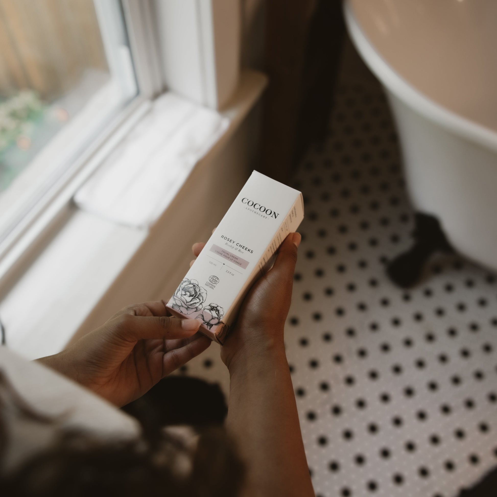 Person holding a box of Rosey Cheeks Facial Cream in a bathroom setting