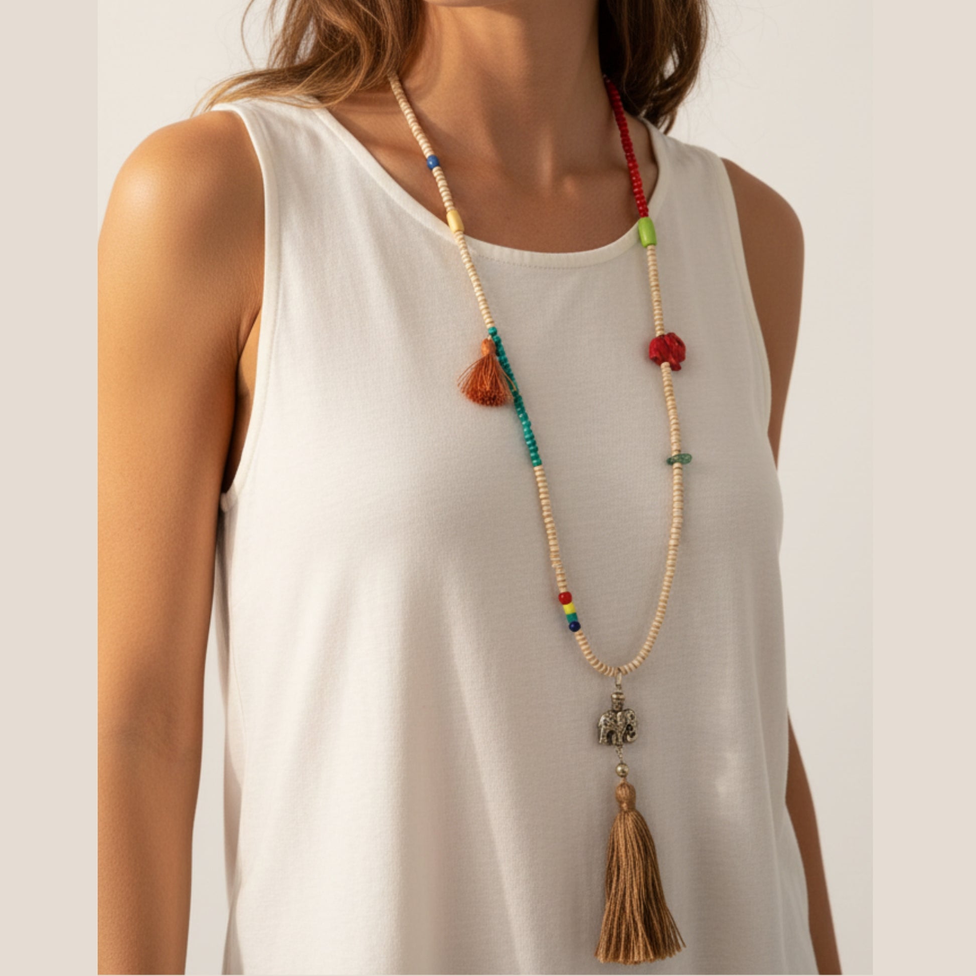 Long colorful mala necklace with cream seed beads, multicolor accents, mini tan tassel, red fabric elephant charm, antique silver elephant pendant and large tan tassel drop worn by a woman — handmade in India for boho and spiritual style