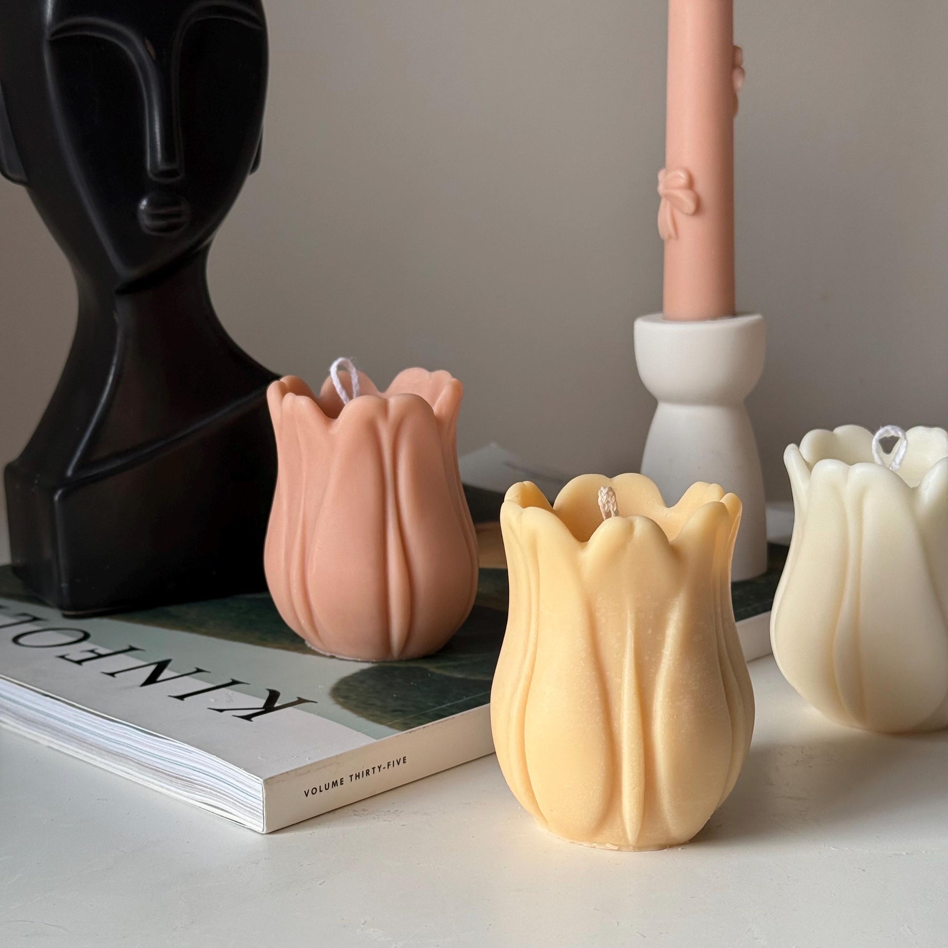 Decorative tulip-shaped candles on a surface with a book and abstract sculpture in the background.