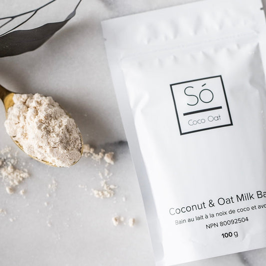 Spoonful of coconut and oat milk bath powder next to a package labeled 'Sö Coco Oat' on a light background.