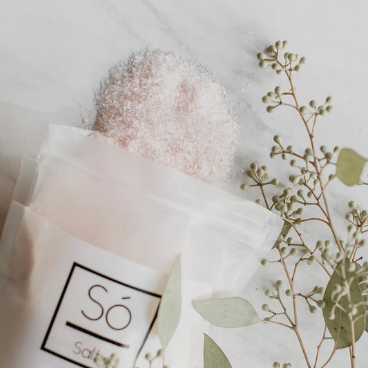 Sack of Só Salty Bath Salts spilling onto a marble countertop with a plant garnish