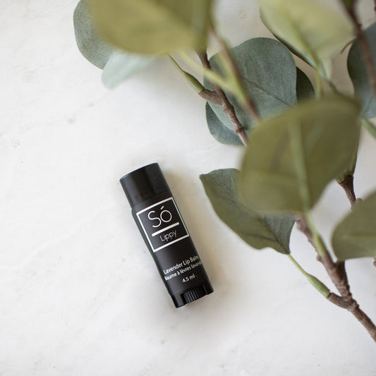Lavender lip balm container with 'Sö Lippy' branding on a white surface with green leaves.