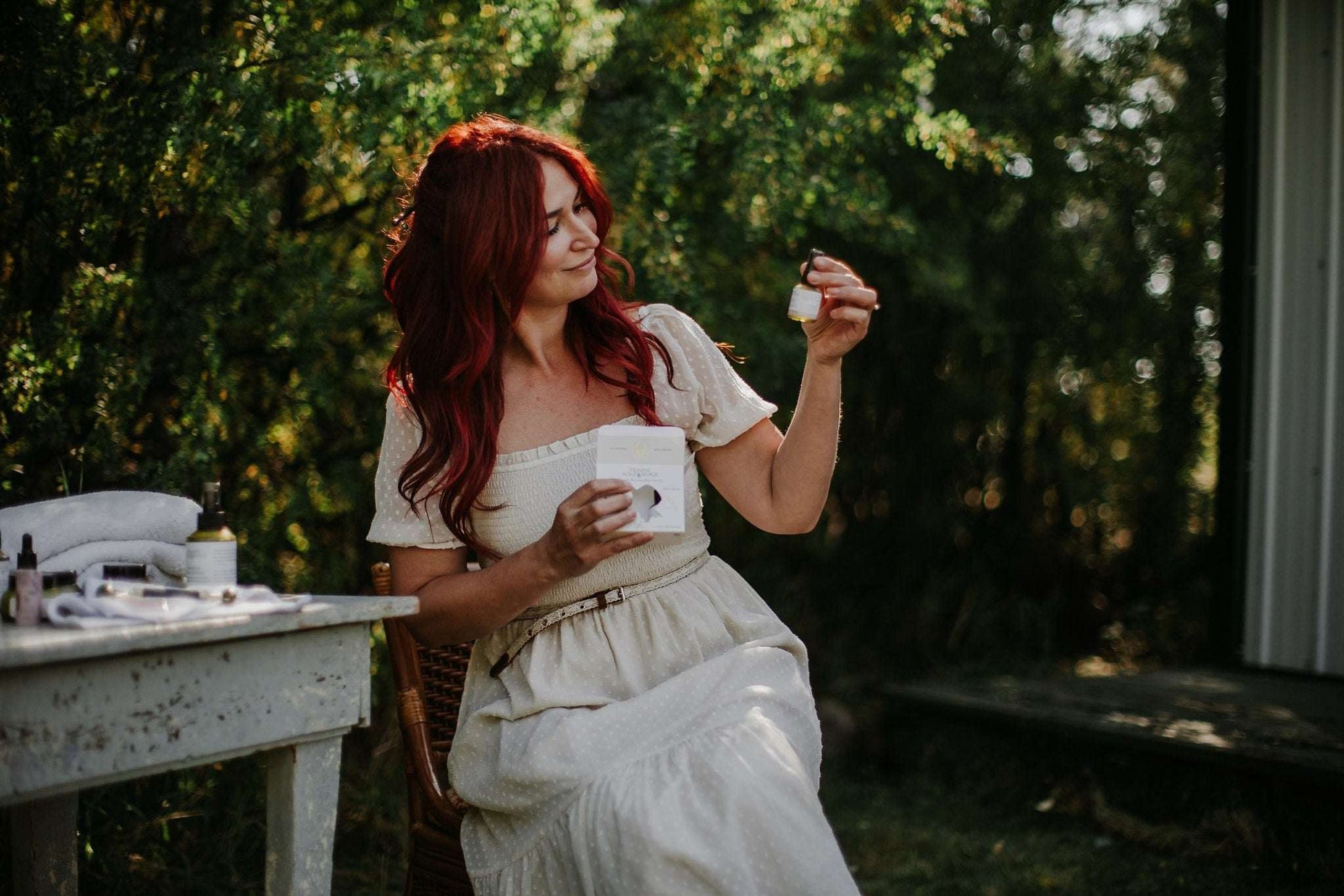 Woman in a white dress sitting outdoors holding a bottle of Mini Skincare Trio.