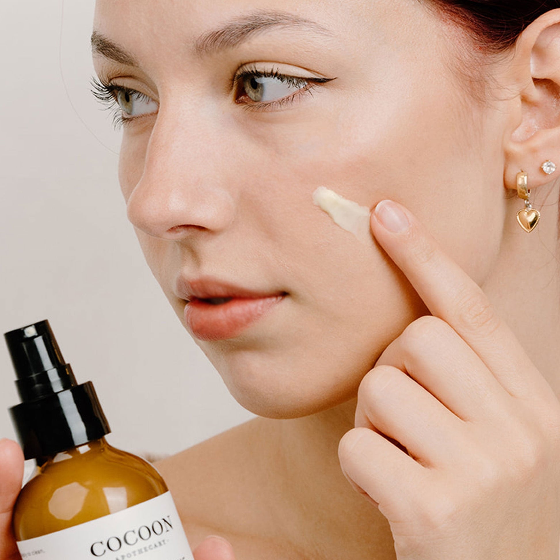 Woman applying Rosey Cheeks Facial Cream to her face with a bottle labeled 'COCOON' in the foreground.