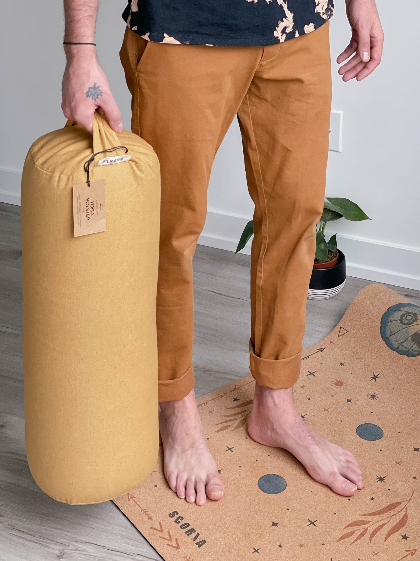 Person holding a mustard-colored yoga bolster on a wooden floor with a plant in the background