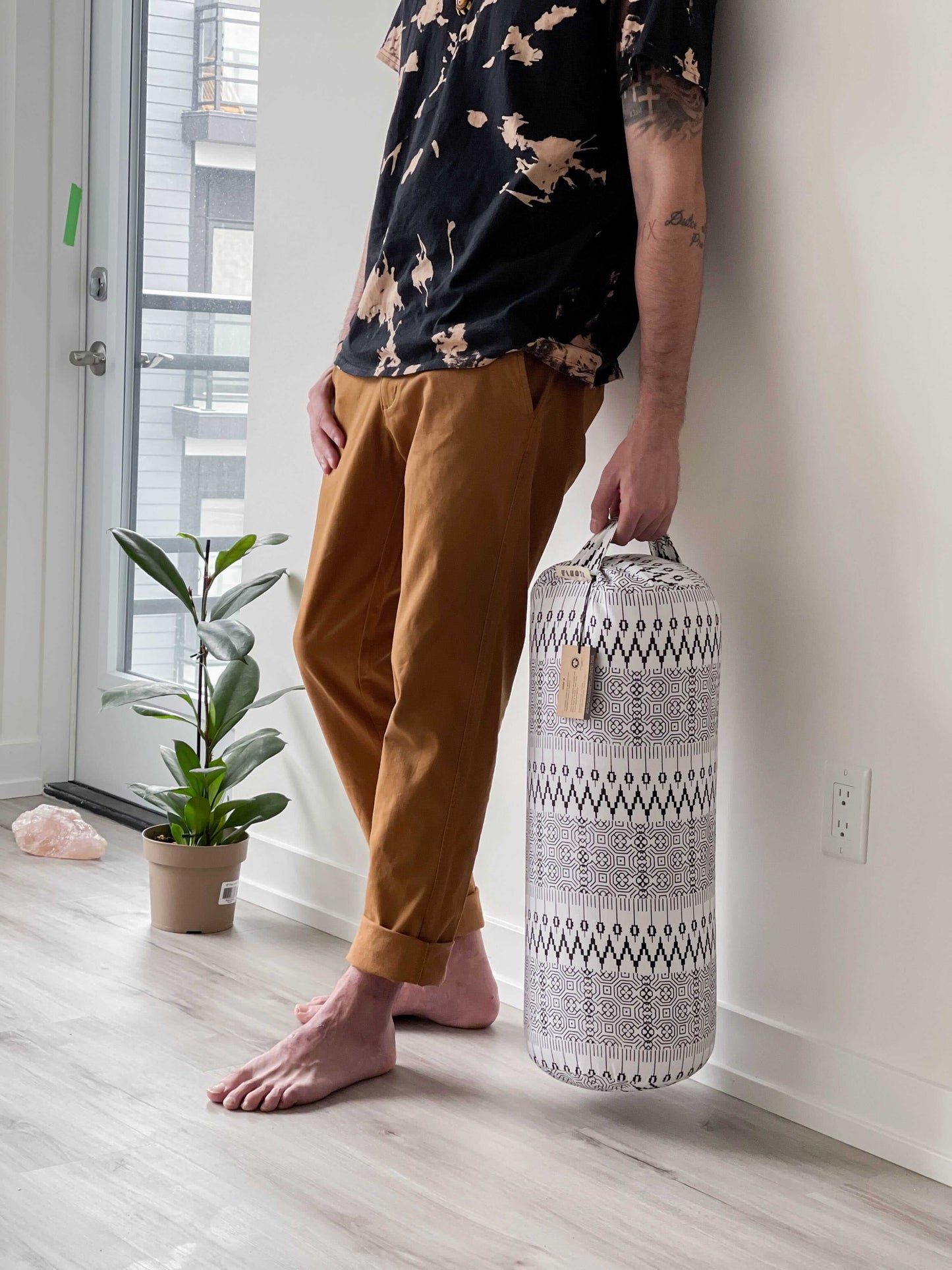 Person holding a patterned yoga bolster in a room with a plant and window.