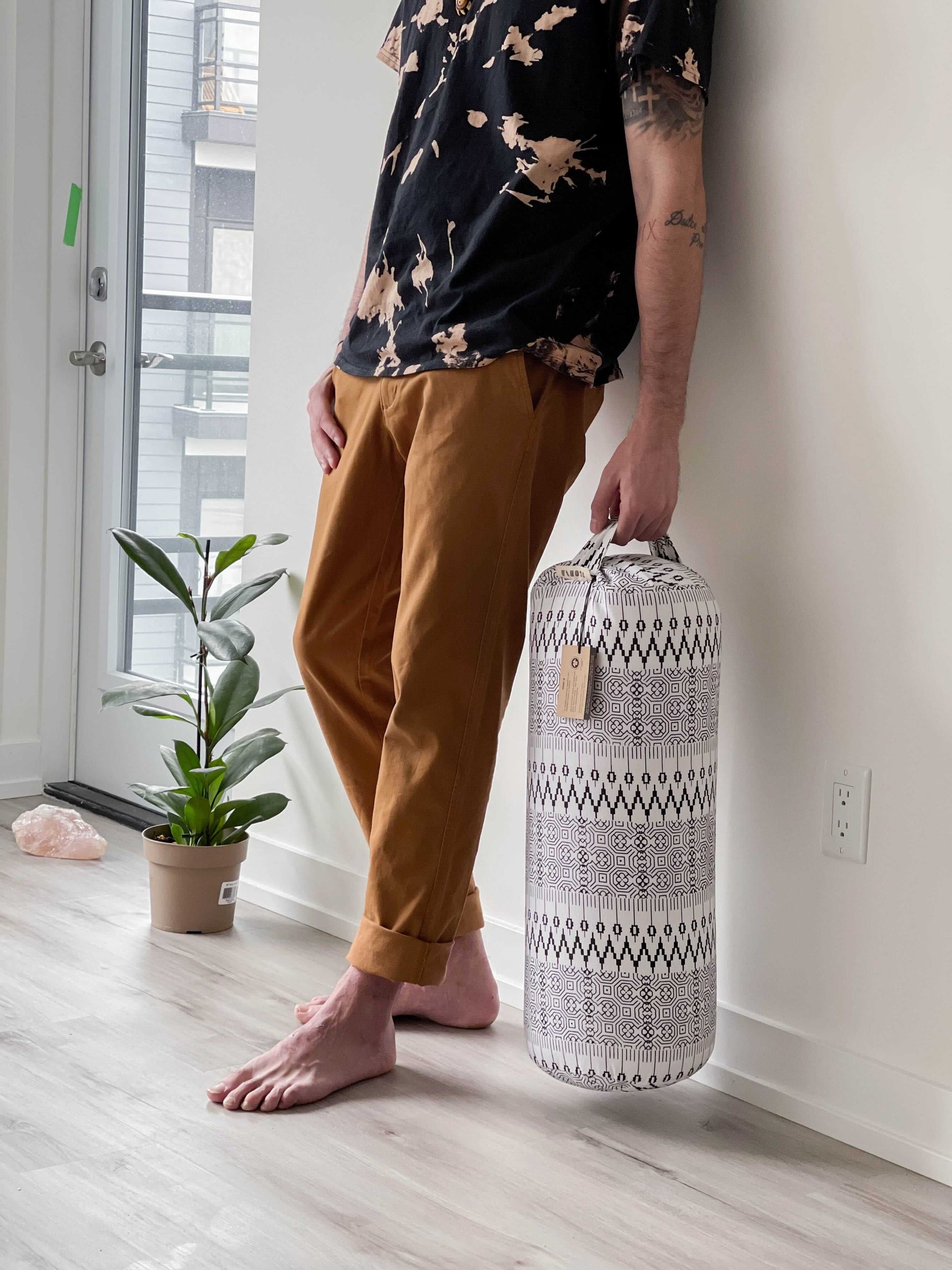 Person holding a patterned yoga bolster in a room with a plant and window.