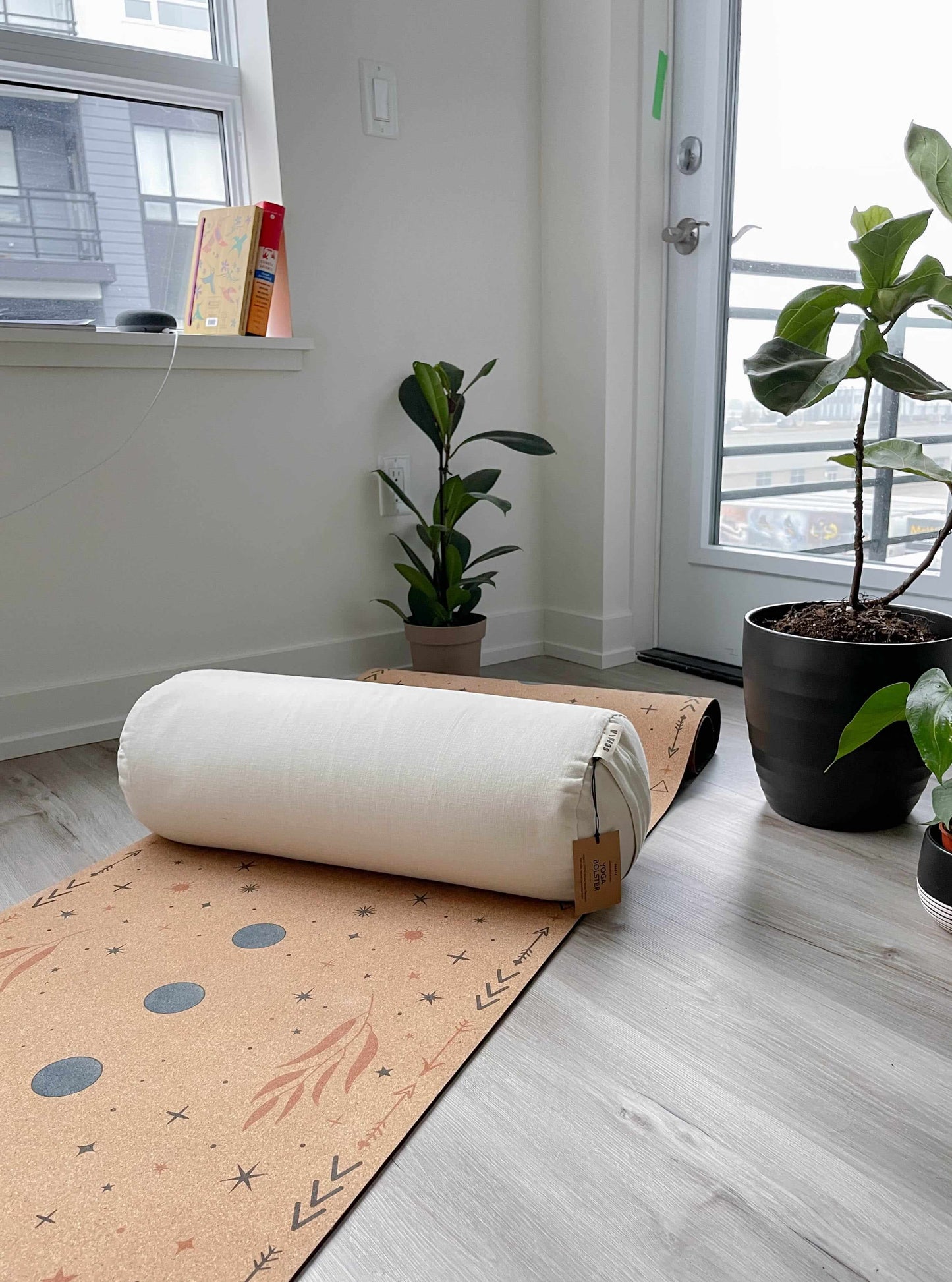 White cylindrical bolster pillow on a cork yoga mat on a wooden floor with plants and a window in the background
