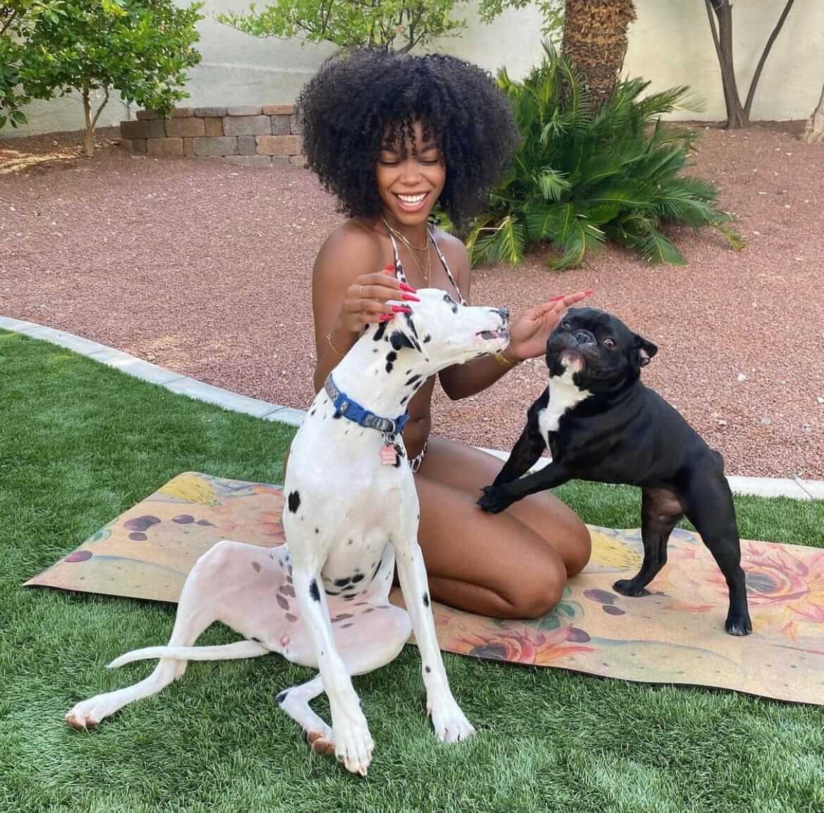 Woman sitting on a cork yoga mat with two dogs, one Dalmatian and one black dog, outdoors.