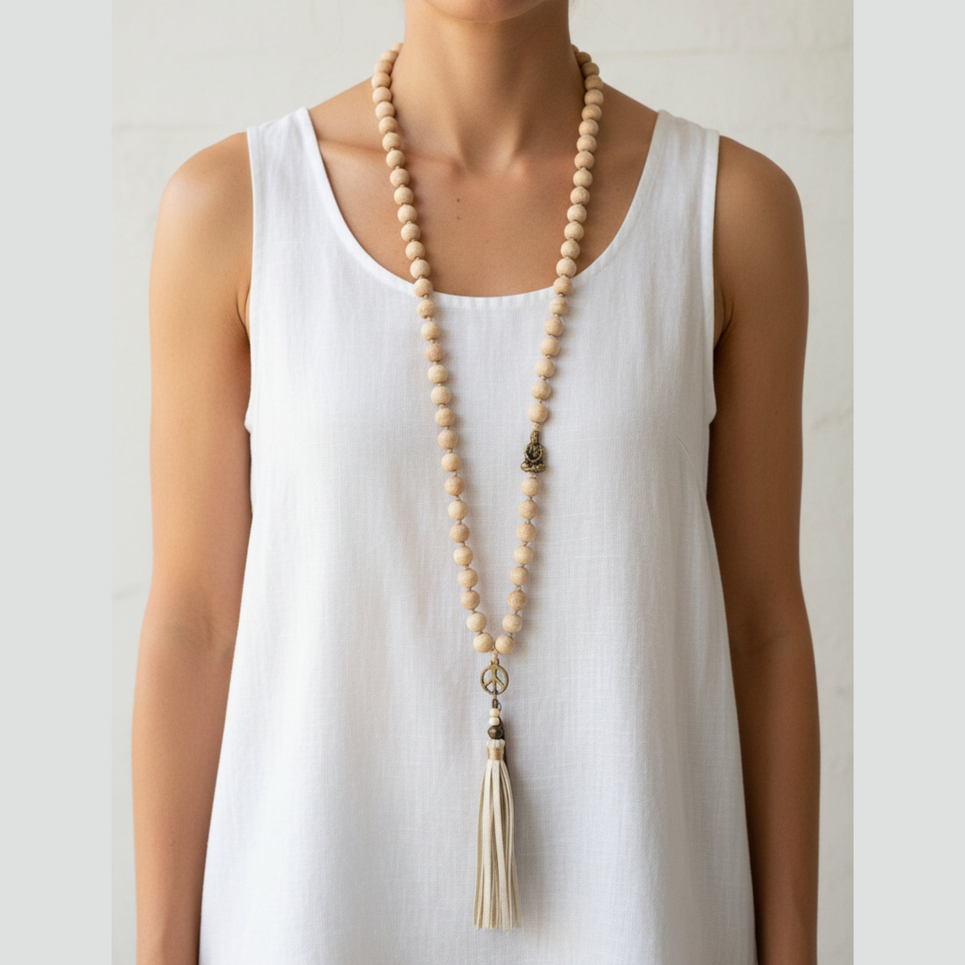 Long natural wood bead mala necklace with antique gold Buddha charm, peace sign pendant and cream leather tassel worn by a woman — handmade in India for boho, yoga, and spiritual style