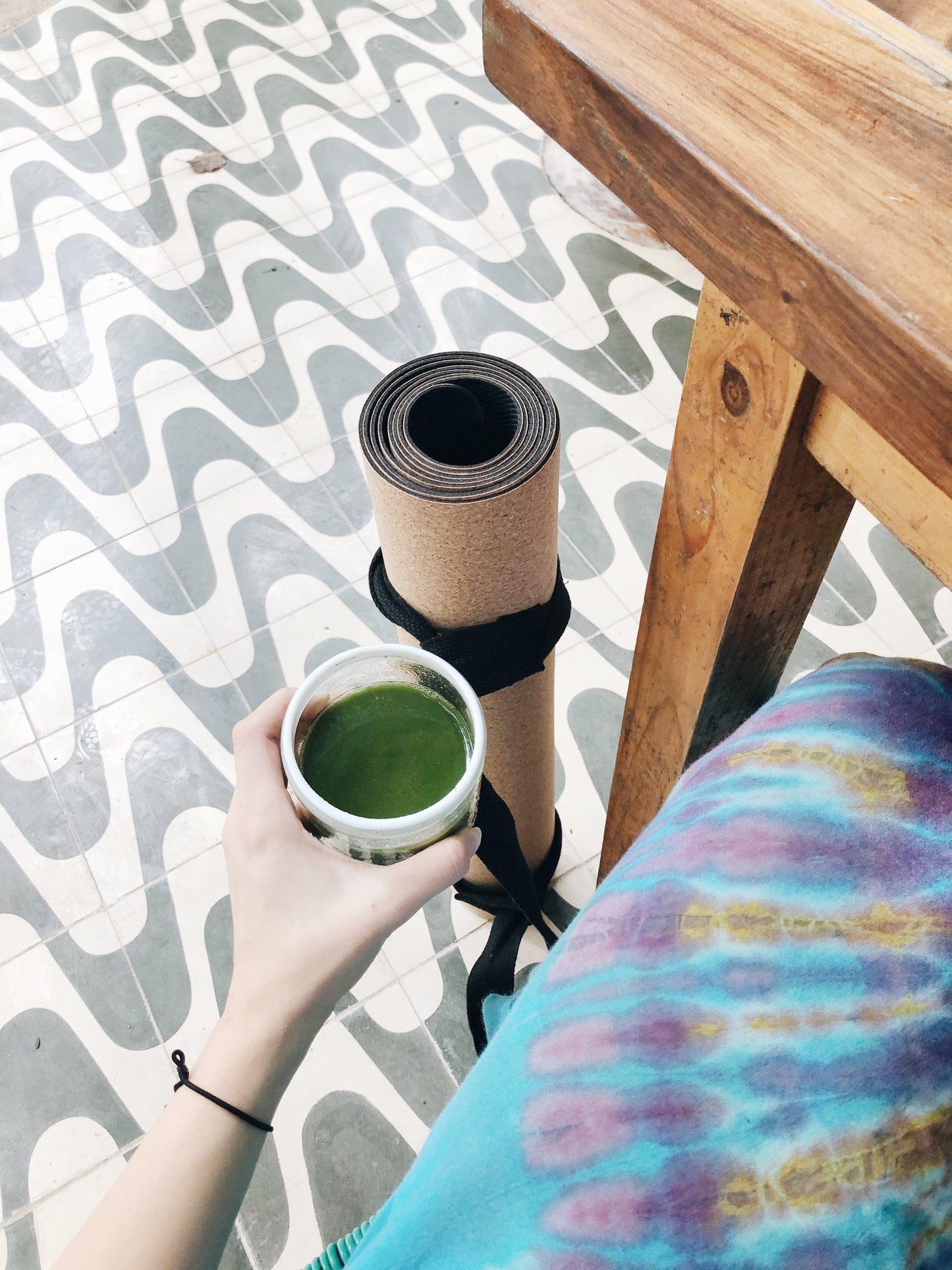 Person holding a cup of green tea next to a yoga mat on a patterned floor.