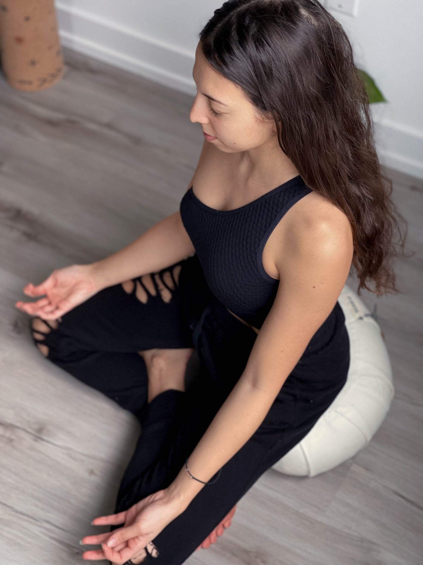 Woman in black outfit sitting on a white meditation cushion on a wooden floor.