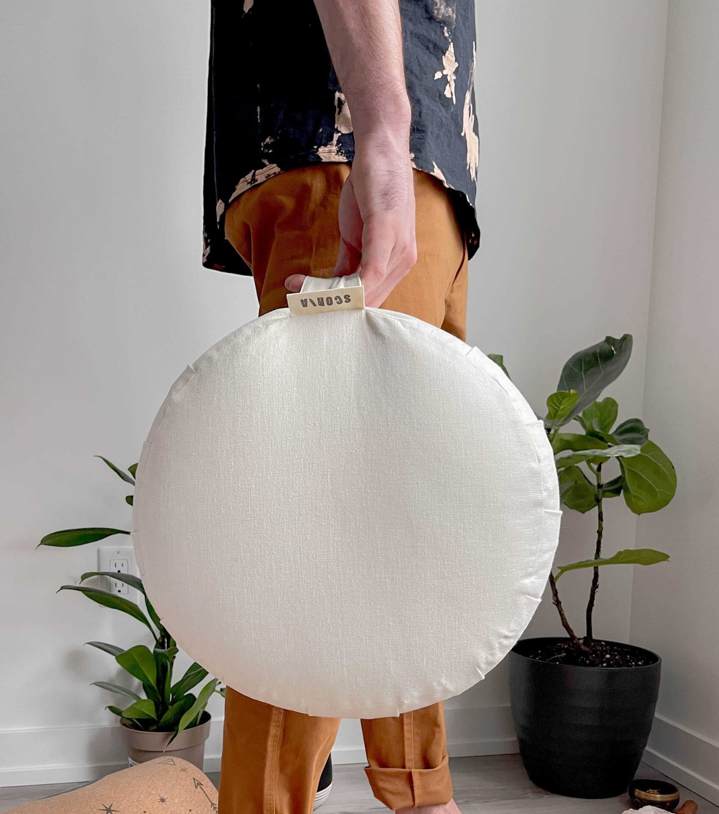 Person holding a round white cushion with a brand tag, standing in a room with plants.