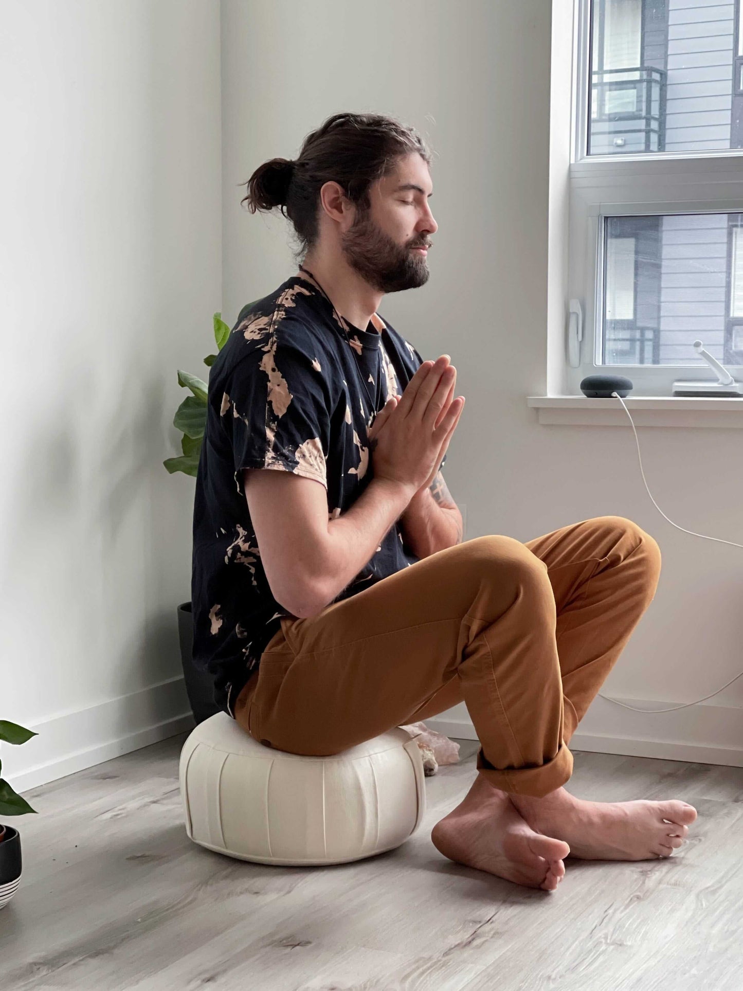 Man meditating on a cushion in a room with a window and plant